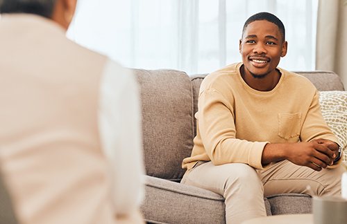 A smiling man sits on a sofa speaking with a therapist