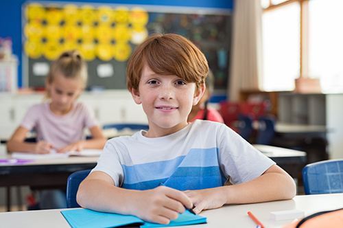 Elementry school boy with red hair sits in a classroom smiling at the camera